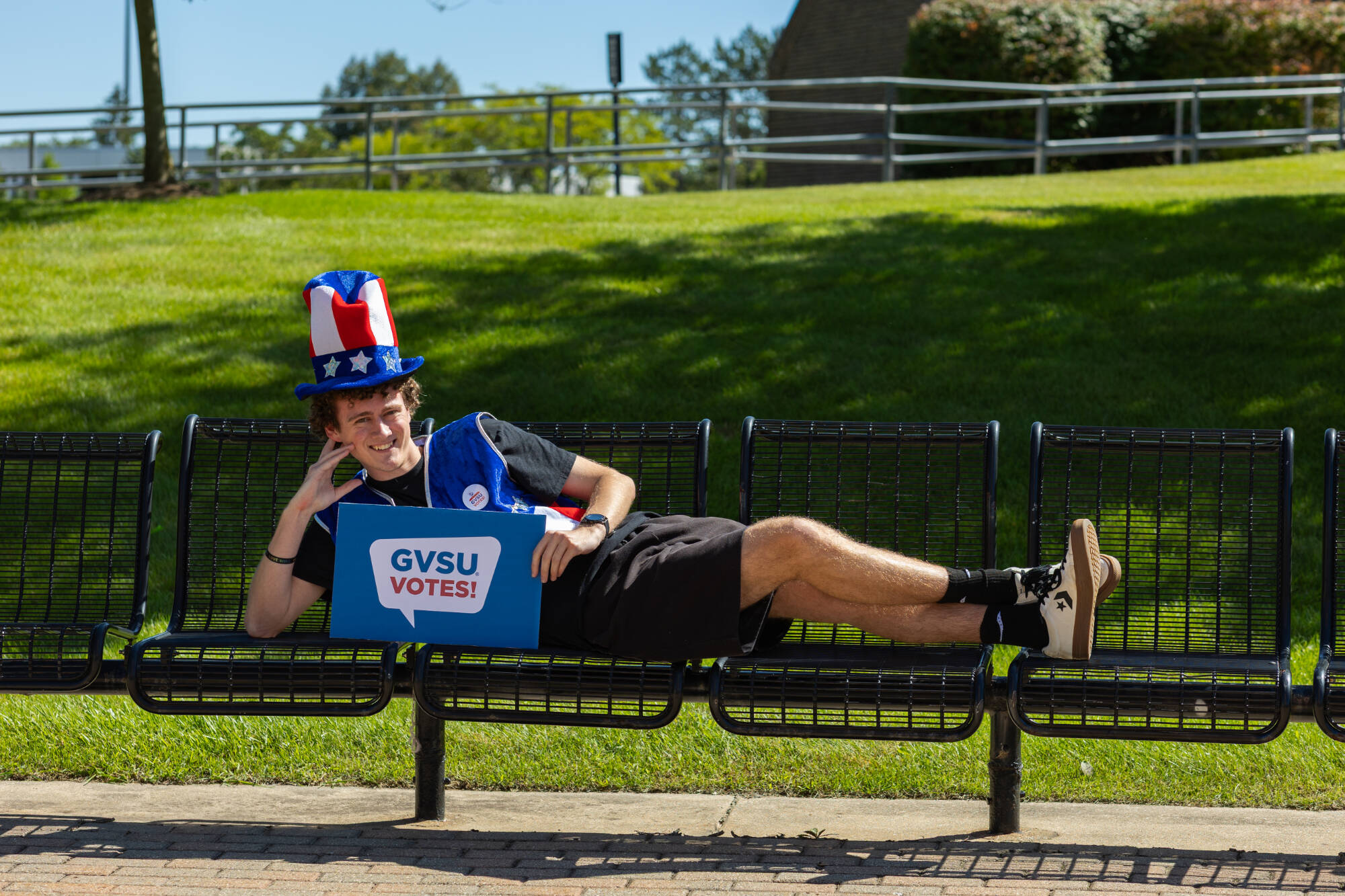 A student on a bench wearing an Uncle Sam hat and holding a sign that says "GVSU Votes!"